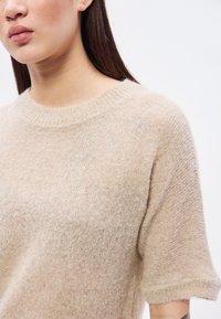 Šviesiai smėlio spalvos megztinis trumpomis rankovėmis, apvalia iškirpte ir švelnia tekstūra, turintis šiek tiek permatomą išvaizdą.