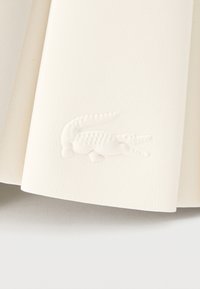 Λευκό υλικό με υφή, με ανάγλυφο λογότυπο κροκόδειλου κοντά στην άκρη, πάνω σε μια ανοιχτόχρωμη, λεία επιφάνεια.