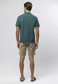 Tumši zaļa īsroku polo krekls ar teksturētu audumu, kombinēts ar gaiši bēšām ceļgalu garuma šortiem un brūnām čībām. Skats no muguras.