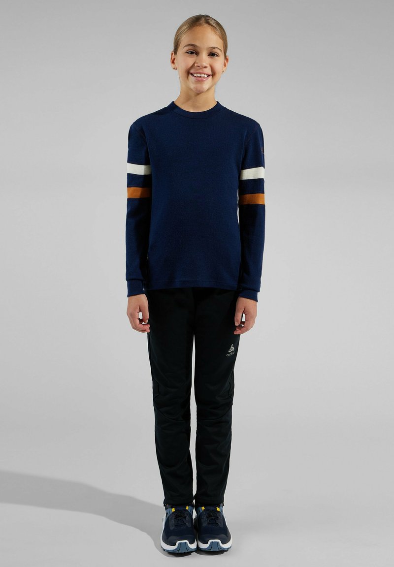 Marineblauer Langarmpullover mit cremefarbenen und braunen Streifen, kombiniert mit schwarzen Sporthosen. Die Sneaker haben Akzente in Blau, Grau und Gelb.
