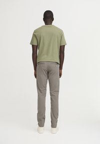 T-shirt di cotone verde oliva abbinata a pantaloni di cotone grigio chiaro, con due tasche posteriori con decorazioni dorate e sneakers bianche.
