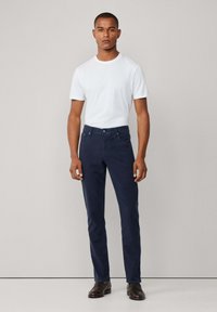 Witte crew-neck t-shirt, getailleerd. Donkerblauwe broek, rechte pijpen, katoenmix met twee voorzakken en knoopsluiting, gecombineerd met donkere schoenen.