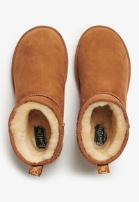 Botas de tobillo de ante color marrón con forro de piel de borrego y un borde decorativo cosido. Presentan una etiqueta de tirón con la marca para facilitar su colocación.
