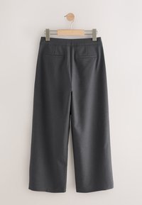 Pantalon large gris foncé suspendu sur un cintre en bois contre un mur blanc uni, vu de dos avec deux poches passepoilées.