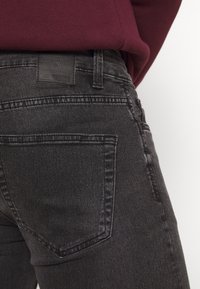 Schwarze Denim-Jeans mit körpernahem Schnitt, Ausstattung mit Gesäßtaschen und einem Markenlederpatch an der Taille. Die Textur wirkt glatt.