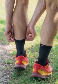 Calcetines tobilleros negros con textura de malla y un pequeño logotipo de color, combinados con zapatillas deportivas rojas con acentos amarillos y una suela resistente.