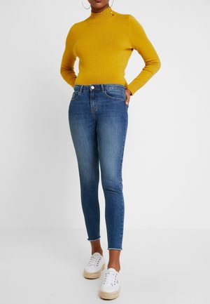 Persoon die een mosterdgele trui met lange mouwen draagt en een blauwe skinny jeans met een hoge taille, gecombineerd met witte plateau sneakers.