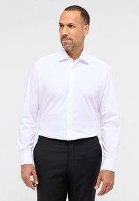 Weißes Langarm-Hemdkleid mit Kragen, Knopfleiste und glatter Textur, kombiniert mit schwarzen Hosen. Taillierter Schnitt betont die Silhouette.