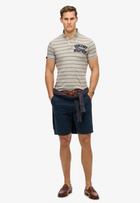 Grå stribet polo t-shirt med krave, marineblå shorts og brune loafers. En marineblå sweater er bundet om livet.
