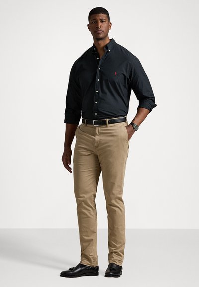 Svart skjorta med knappar, khakifärgade chinos, svart bälte, svarta loafers. Skjortan har krage och broderad logotyp; byxorna har slim fit.