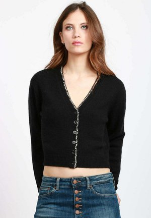 Cardigan corto nero con scollo a V, chiusura con bottoni e bordo testurizzato a contrasto lungo il colletto. Indossato sopra un paio di jeans.