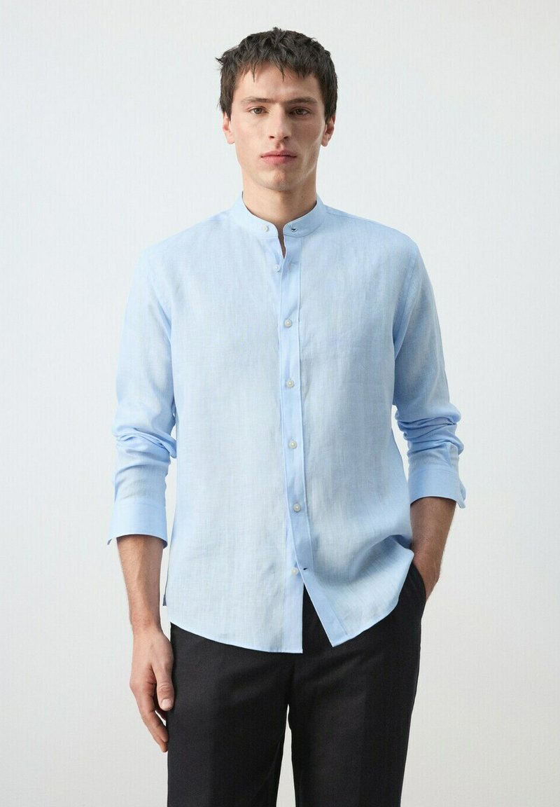 Homme portant une chemise bleu clair à col mandarin et un pantalon noir, debout avec une main dans la poche, sur fond blanc.