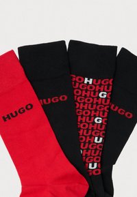 Štyri páry ponožiek: jeden červený s veľkým čiernym nápisom "HUGO", dva čierne s opakujúcim sa "HUGO" v červenej a bielej farbe, vyrobené z bavlnenej zmesi, s rebrovanou textúrou.