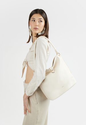 Giovane donna con abbigliamento beige e grande borsa a tracolla bianca, guarda di lato su sfondo semplice.