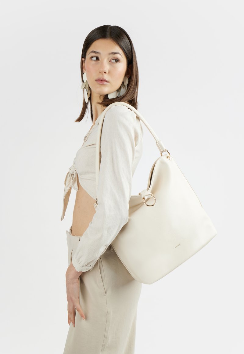 Young woman wearing beige outfit and large white shoulder bag, looking sideways against plain background.
