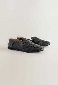 Pair of black leather loafers with a low heel and a strap detail on a light neutral background.