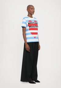 Blå og hvit stripet t-skjorte med et stort rødt hjerte og "MOSCHINO"-tekst i svart. Kombinert med svarte vide bukser og spisse sko.