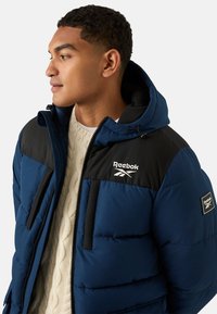 Giacca a vento blu con dettagli neri, tasche con cerniera e logo Reebok. Caratterizzata da cappuccio, tessuto strutturato e design isolante.