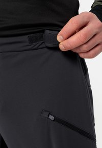 Pantalons de sport noirs avec une texture lisse, dotés d'une fermeture Velcro à la taille et d'une poche zippée sur le côté.