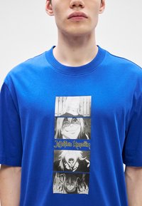 Μπλε βαμβακερό t-shirt με γραφική εκτύπωση τεσσάρων πάνελ anime χαρακτήρων και κείμενο "JUJUTSU KAISEN" σε κίτρινο από κάτω. Στρογγυλή λαιμόκοψη.