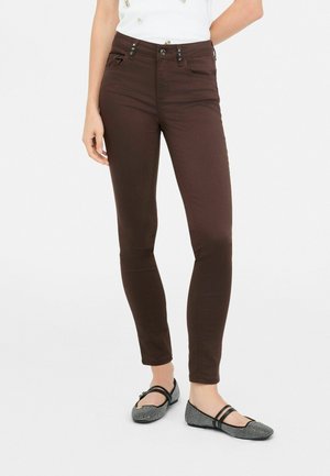 Pantaloni slim-fit marroni con vita alta, due tasche anteriori con bottoni decorativi e una texture liscia. Abbinati a ballerine nere con stampa.