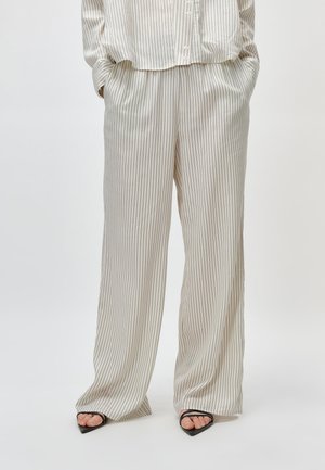 Osoba ubrana w luźne kremowe spodnie w pionowe paski z kieszeniami oraz czarne sandały na obcasie z odkrytymi palcami, stojąca na jednolitym tle.