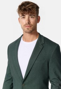 Waldgrüner Fischgrätenblazer mit einem einzelnen Knopfverschluss, Reverskragen und einer schlanken, taillierten Passform über einem weißen T-Shirt.