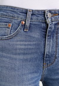 Jeans de mezclilla azul claro con un diseño clásico de cinco bolsillos, remaches de cobre, costuras en contraste y detalles de desgaste en la tela.