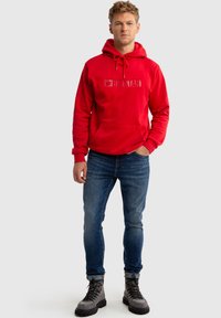 Raudona medvilninė hoodie su kengūros kišene ir įspaustu "BIG STAR" logotipu. Dėvima su mėlynomis aptemptomis džinsomis ir pilkais bateliais su raišteliais.