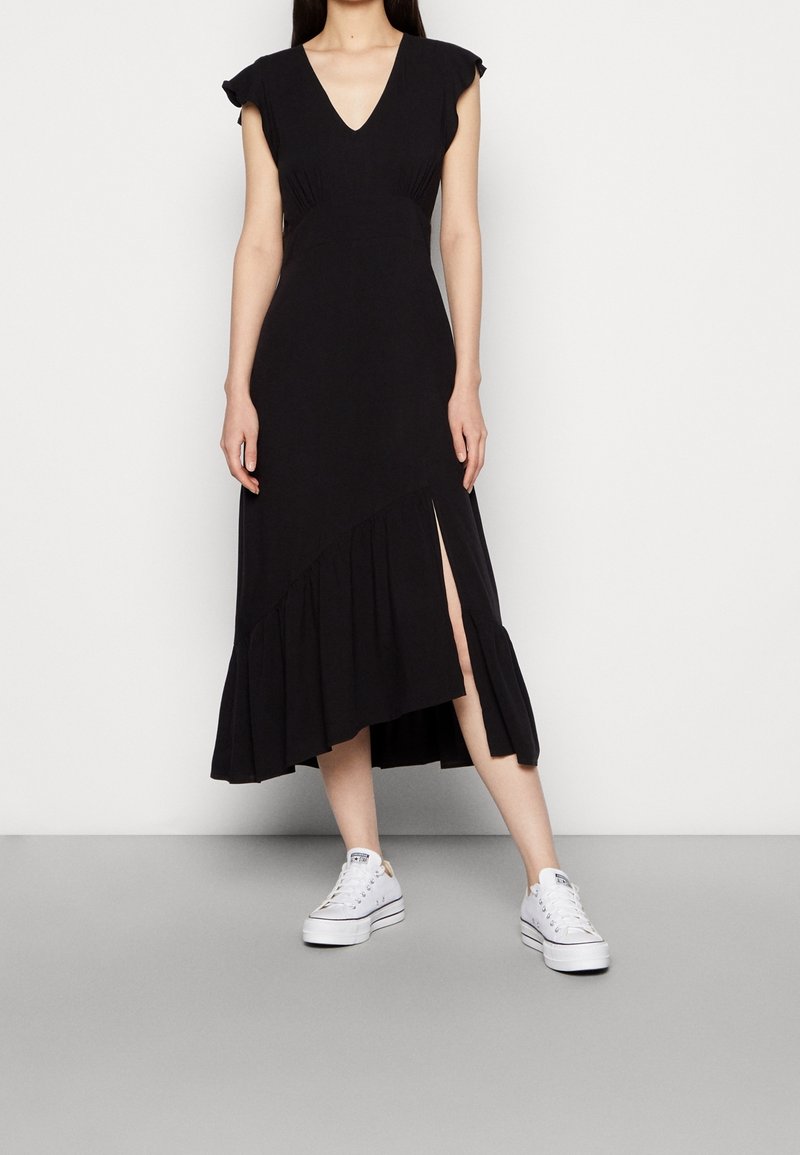 Femme portant une robe midi noire sans manches avec ourlet à volants et fente avant, associée à des baskets basses blanches, debout sur un sol gris.