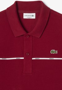 Bordeaux polo shirt van textiel met een gestructureerde stof, met een knoopsluiting van twee knopen, witte horizontale strepen met het opschrift "LACOSTE" en een groene krokodil logo op de borst.