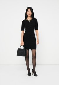 Femme portant une robe noire texturée avec un col en chaîne dorée, des collants noirs transparents, des talons hauts et tenant un sac à main noir avec des clous.