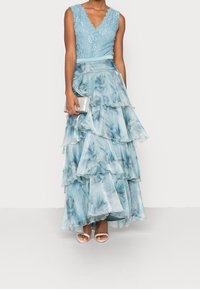 Robe bleue claire avec un corsage en dentelle florale à décolleté en V, jupes à volants superposés dans des nuances variées de bleu, associée à une pochette argentée et des chaussures ouvertes.