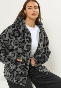 Femme portant un sweat à capuche zippé en peluche gris et noir à motif léopard, avec une chemise blanche et un pantalon noir, assise les mains dans les poches.