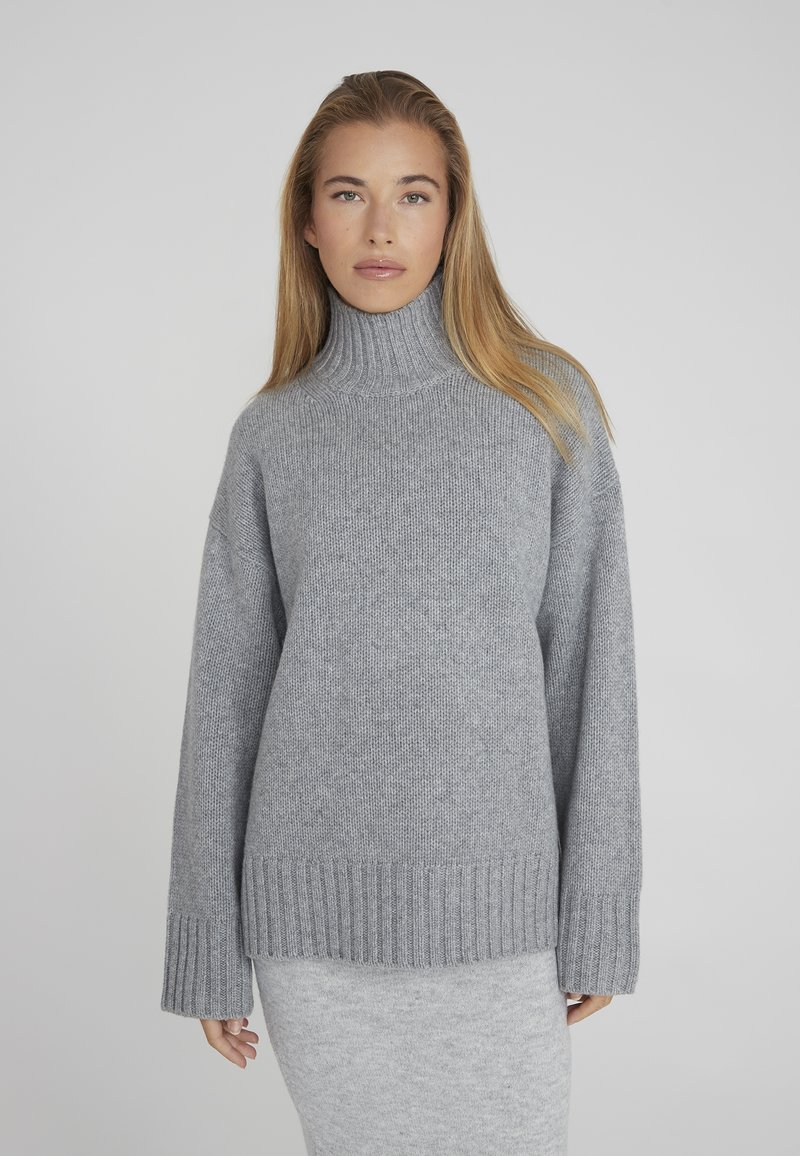 Pull en col roulé gris avec un tricot texturé, poignets et ourlet côtelés. Coupe décontractée avec épaules tombantes, porté sur une jupe gris clair.