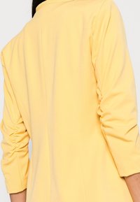 Veste jaune légère à manches trois-quarts, avec des détails froncés aux épaules et un tissu lisse légèrement texturé.