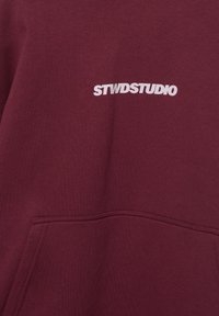 Bordeauxrode hoodie gemaakt van zachte stof, voorzien van een zak aan de voorkant en een wit "STWDSTUDIO" logo. Gladde textuur met stikdetails.