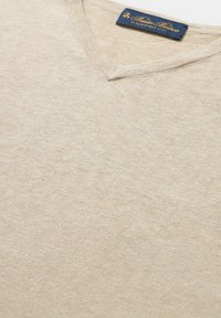 Beiger V-Ausschnitt-Pullover aus weichem, glatten Stoff. Verfügt über eine dezente Textur und ein marineblaues Etikett am Hals mit Markendetails.