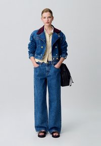 Chaqueta de mezclilla con un cuello burdeos, camisa de rayas amarillas debajo, jeans azules de pierna ancha y bolso negro. Sandalias con un acento dorado.