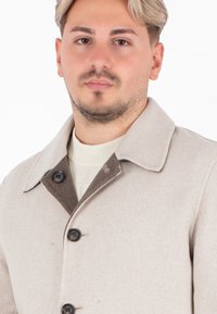 Cappotto in misto lana beige con colletto marrone, dotato di due bottoni e tasche frontali. Texture liscia, dettagli di cucitura minimi, design classico.