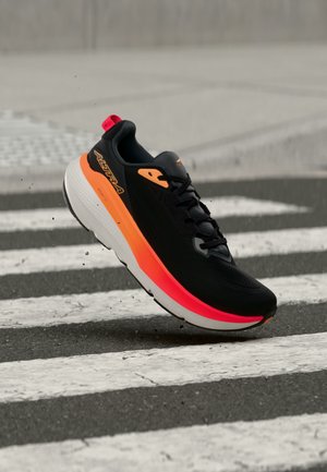Chaussure de course noire et orange avec semelle blanche épaisse flottant au-dessus d'un passage piéton sur chaussée en asphalte.
