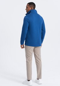 Chaqueta de forro polar azul con un alto cuello, aberturas laterales y una textura suave, combinada con pantalones beige claros y zapatillas blancas.