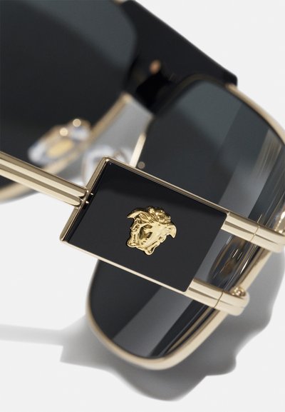 Las gafas de sol con detalles en oro presentan un grueso marco negro, grandes lentes grises y un prominente emblema de Medusa dorado en la patilla.