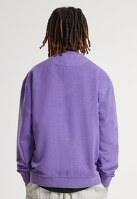 Felpa viola con una vestibilità rilassata, polsini e bordo a costine. Il materiale appare morbido con una finitura leggermente testurizzata. Vista posteriore mostrata.