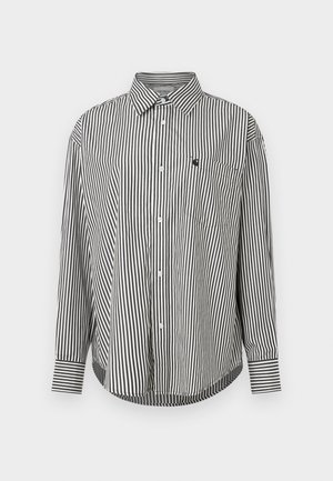 Črna in bela vertikalno prugasta srajca z dolgimi rokavi, s koničastim ovratnikom, sprednjimi gumbi in žepom na levi strani prsnega dela. Ima kontrastne manšete.