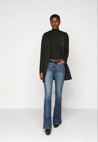 Top preto de manga longa sob uma jaqueta com capuz verde-escuro, combinado com jeans azul de perna larga e sapatos pretos de salto. Texturas suaves e ajustadas.