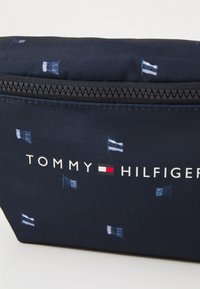 Laatikko sininen kangaspussi, jossa on vetoketjukiinnitys, ja siinä on toistuva kuvio lippuja ja teksti "TOMMY HILFIGER" valkoisilla ja punaisilla yksityiskohdilla.