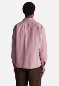 Jasnoróżowa, długi rękaw koszulka wykonana z bawełny, z zapięciem na guziki, bocznymi rozporkami i luźnym krojem, pokazana z tyłu.