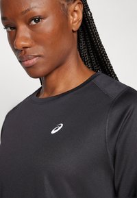 Primo piano di una donna con capelli intrecciati che indossa una maglietta sportiva nera con il logo bianco ASICS sul petto.