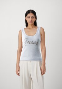 Camiseta sin mangas blanca con textura acanalada que presenta un logo metálico plateado "Guess" en el pecho; escote redondeado y tirantes anchos.